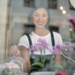 Smiling young Asian waitress female in apron looking at camera while standing behind glass window with beautiful flowers on windowsill in coffee shop and holding clear glass lid over dessert on cake stand