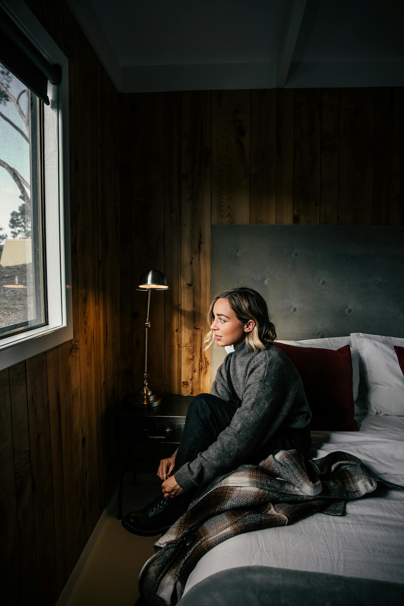 Young woman sitting by the window in a cozy bedroom, lost in thought. Warm and peaceful atmosphere.